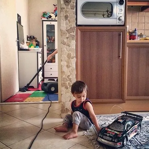 A boy plays with his toy car as his mother cleans the flo
